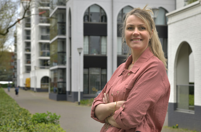 Anneke Luijten, blond haar, roze blouse, poseert met armen over elkaar voor appartementengebouw in Tilburg