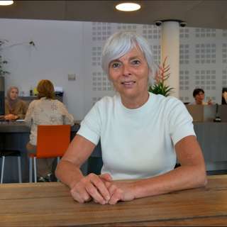 Caroline Timmermans zit aan tafel in woonwinkel Tilburg met mensen op de achtergrond en kijkt in de camera